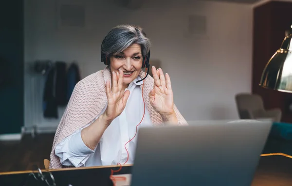 Happy middle aged female in casual clothes and headset smiling and looking at computer screen while sitting at table with laptop