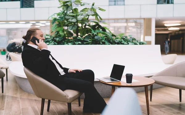 Side view of male entrepreneur in formal wear sitting on soft chair in executive lounge with plant while talking on mobile phone and working on laptop