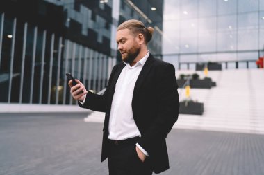 Siyah takım elbiseli ciddi sakallı bir işadamının yan görüntüsü. Sokakta durup eşlerini beklerken cep telefonunu karıştırıyor.