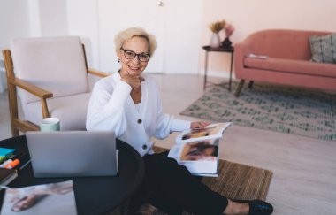Gözlüklü, gülümseyen, olgun bir iş kadını. Yuvarlak ahşap masada dizüstü bilgisayar ve dergi okuyarak modern oturma odasında kameraya bakıyor.