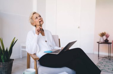 Düşünceli orta yaşlı serbest çalışan bir kadın dizüstü bilgisayarla masada oturuyor ve uzaktan kumanda sırasında kalemle dudaklara dokunurken gözlerini kaçırıyor.