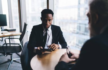 Afrika kökenli Amerikalı erkek girişimci panoramik pencerelerin yanındaki masada oturuyor ofisteki meslektaşıyla konuşurken telefona bakıyor.