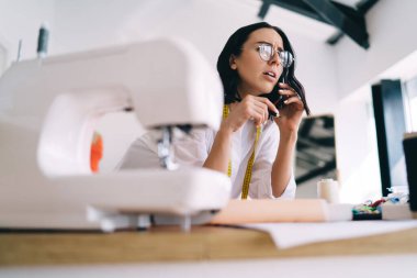 Cep telefonuyla konuşan ve dikiş makinesinin yanında duran kafası karışık kadın terzinin seçici odak noktası.