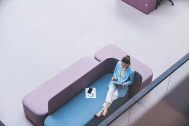 Mavi takım elbiseli tam vücut yüksek açılı genç bayan işçi rahat bir kanepe ve tablet otururken modern dinlenme odasında cam duvarla dinleniyor.