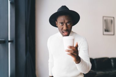 Siyah şapkalı ve beyaz balıkçı yakalı ciddi bir Afro-Amerikalı adam elinde kağıt bardak sıcak içecekle ışık odasında dururken kameraya hoşnutsuzca bakıyor.