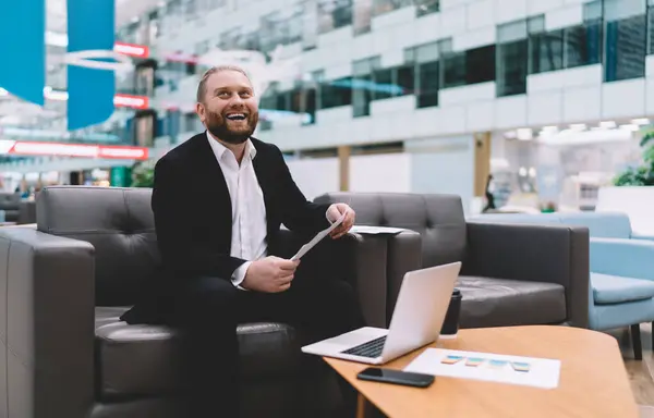 Successful businessman excited to receive good news in documents while sitting at table with laptop smartphone coffee cup and smiling while looking up at ceiling in blurred spacious hall