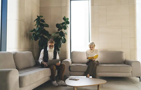 Full body of elderly coworkers relaxing on sofa while female reading interesting book and male looking at screen of laptop while browsing near table with notepad in daytime in reception room of office
