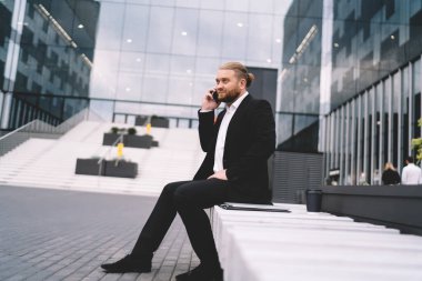 Sakallı ciddi girişimci dış ofis terasında oturmuş elinde not defteri ve kahveyle akıllı telefondan konuşurken başka tarafa bakıyor.