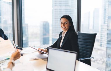 Resmi olarak kadın yöneticinin kimliği belirsiz iş arkadaşlarıyla iş konularını konuşurken ofis masasında oturması ve kameraya bakması.