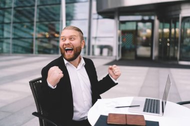 Resmi olarak mutlu sakallı genç girişimci başarılı iş projesini kutlarken dizüstü bilgisayar ve not defteriyle aynı masada oturuyor.