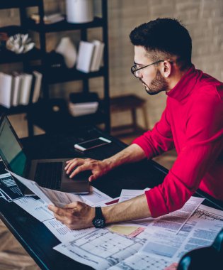 Konsantre erkek, kağıt belgeler ve modern dizüstü bilgisayarla uzaktan yakından ilgilenen modern bir apartman dairesinde çalışan ciddi hippi adam web sitesinde bilgi kontrol ediyor.