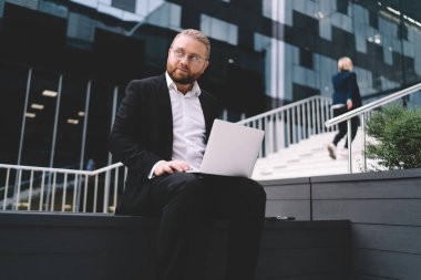 Seçkin takım elbiseli ve gözlüklü genç, sakallı bir girişimci bankta oturup başka tarafa bakarken netbook üzerinde çalışıyor.