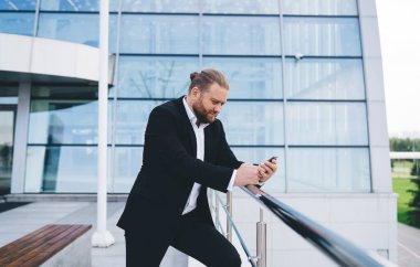 Kentteki modern ofis binasına karşı tırabzanda duran ve cep telefonuna göz atan siyah takım elbiseli genç işadamının yan görüntüsü
