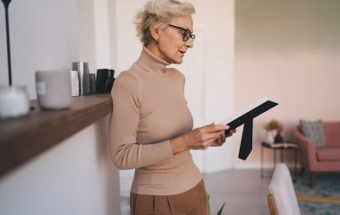 Gayri resmi çerçevede tutan ve evdeki yerli insanların resimlerini düşünen, günlük giysili ve gözlüklü hoş yaşlı bayanın yan görüntüsü.