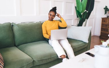 Akıllı telefonlu kanepenin yanındaki koltukta oturan ve oturma odasındaki görüntülü sohbet sırasında bilgisayarın ekranına bakan Afro-Amerikan kadın.