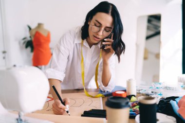 Çalışan kadın moda tasarımcısı günlük giysiler ve göz çizimi desenleri giyer ve çalışma alanında santimetre bandıyla ayakta dururken cep telefonuyla sohbet eder.