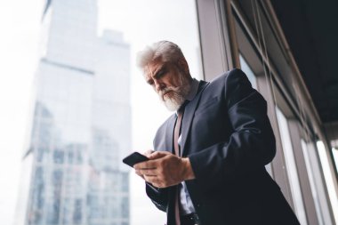 Ciddi gri saçlı, takım elbiseli bir girişimci şehrin modern iş merkezinde çalışırken pencerenin yanında durup cep telefonu kullanıyor.