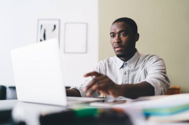 Kendinden emin Afrikalı Amerikalı erkek serbest çalışan bulanık kırtasiye malzemeleriyle masada oturuyor ve evde uzaktan çalıştırma için yazarken ve çalışırken bilgisayarın ekranına bakıyor.