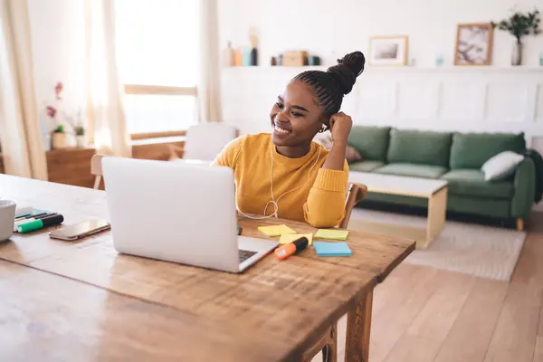 Smiling young black female freelancer with braided hair sitting at table with smartphone and listening to conversation in earphones while looking at screen of laptop and working from home