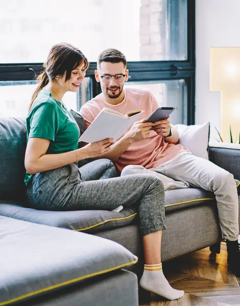 Husband with e-book in hands reading information from literature book in hands of wife resting on couch at home interior, young marriage with touch pad and bestseller spending leisure in apartment