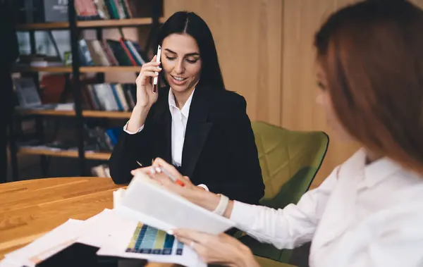 Positive young businesswoman in formal outfit sitting at wooden table and talking on smartphone while discussing project with female coworker in modern office