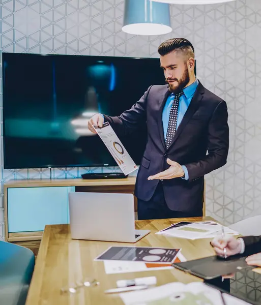 Proud CEO dressed in suit holding paper statistics of financial reports and explaining new strategy of profit to best employee which sitting at meeting table and making notes in notebook in office