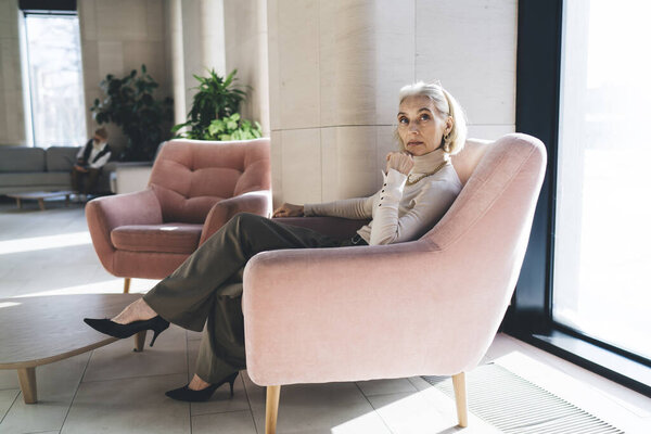 Полный вид со стороны тела задумчивой пожилой женщины в стильном наряде, сидящей на удобном кресле и смотрящей в камеру во время отдыха от работы