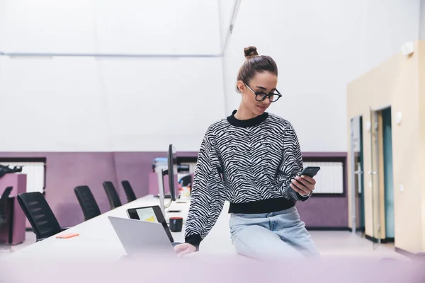 Concentrated young female in casual outfit and eyeglasses sitting on conference table with laptop and browsing mobile phone while working in modern office