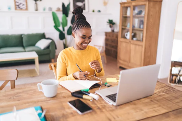 Cheerful young black female student with braised hair taking notes while sitting at table with smartphone markers and looking at screen of laptop with earphones wire at blurred home