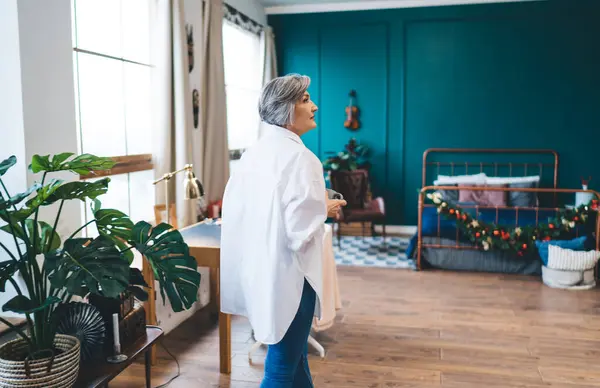 Side view of middle aged female standing near glass window with bright sunlight and green potted plants while looking away in cozy room with bed at home