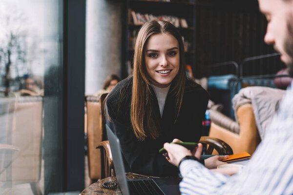 Привлекательная улыбающаяся женщина в повседневной одежде сидит за столом с нетбуком и смотрит в камеру во время встречи с коллегой в кафетерии