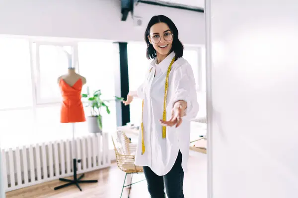 Young smiling designer in white shirt greeting and gesturing to enter workshop with desk and mannequin while looking at camera
