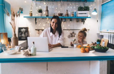 Genç anne, yemek tarifi ararken ve küçük kızıyla yemek pişirirken dizüstü bilgisayarla aynı masada oturuyor.