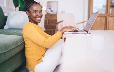 Gözlüklü, gözlüklü, masada dizüstü bilgisayarla oturan ve evde yeni başlayanlar için çalışırken kameraya bakan Afro-Amerikalı kadınların yan görüntüsü.