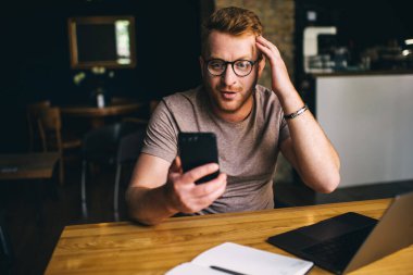 Netbook okuyan etkilenmiş erkek programcı, modern akıllı telefon aygıtı üzerine e-posta aldı. Optik gözlüklü duygusal adam cep telefonu aygıtında gezinen bilgilendirici web yayınıyla şok oldu.