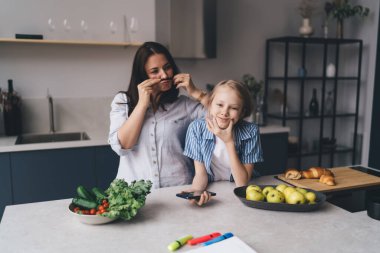 Gündelik giysiler içinde, kızının yanında bıyık takmış, hafta sonu yemek hazırlarken mutfak tezgahında eğlenen neşeli bir anne.