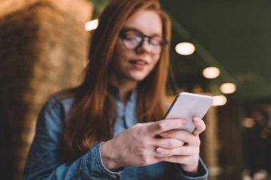 Gündüz vakti kafede otururken akıllı telefondan mesajlaşan günlük kıyafetli ve gözlüklü bir kadının düşük açılı görüntüsü.