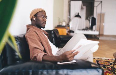 Gözlüklü, kulaklıklı, akıllı telefonlu, kulaklıklı ve gündüzleri başka yere bakarken doküman okuyan düşünceli Afro-Amerikan erkeği.