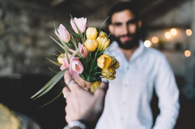 Sakallı, gündelik giysiler içindeki etnik bir adamın odak noktası. İsimsiz bir kadına lale buketini sunarken yatak odasında çelenklerle süslenmişti.