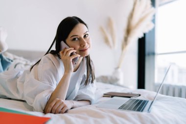 Dizüstü bilgisayarla yatakta yatıp, akıllı telefondan konuşurken rahat kıyafetler içinde gülümseyen ve başka tarafa bakan pozitif genç bir bayan serbest yazar.