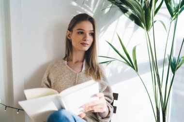 Hafta sonu yeşil bitkinin yanındaki pencere kenarında ilginç kurgu öyküleri okurken başka tarafa bakan ve gülümseyen kadın.