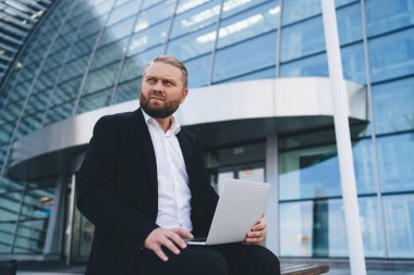 Laptop kullanan ve modern ofis binasına karşı çalışırken yüzünü çeviren resmi giyinmiş genç sakallı adamın alçak açısı.