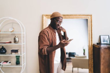 Kapüşonlu Afro-Amerikan erkek cep telefonuna bakıyor ve aynanın yanındaki rahat odada ceketine dokunarak duruyor.