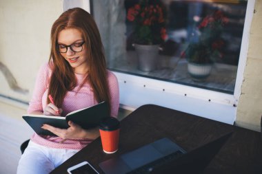 Gözlüklü neşeli, genç bir bayan serbest yazar. Bilgisayarlı, akıllı kahve bardağıyla masada otururken, kalemle aşağıya bakıyor ve yazıyor. Bulanık bitkilerle dolu bir kafenin penceresinin yanında.