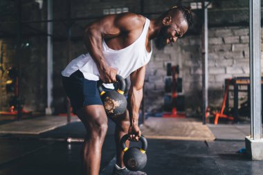 Spor kıyafetli güçlü Afrikalı Amerikalı sakallı erkek sporcunun spor salonunda antrenman yaparken kaslı yüzünü gösteren ağır kettlebell kaldırışının yan görüntüsü.