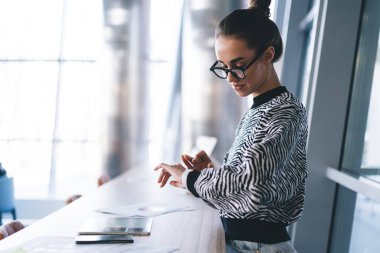 Kol saatini kontrol ederken ve cam duvarla yaratıcı çalışma alanında çalışırken tahta masada tablet ve kağıtla dikilen günlük kıyafetlerle odaklanmış genç bir kadının yan görüntüsü.