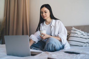 Beyaz gömlekli ve kot pantolonlu genç bir kadın hafta sonu boyunca modern netbook 'a bakarken sıcak içecekle yatakta dinleniyor.