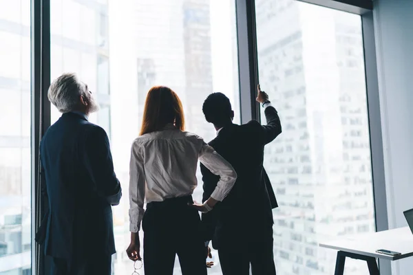 Back view of faceless diverse businesspeople standing near window and observing tall skyscrapers while working in business center and brainstorming startup together