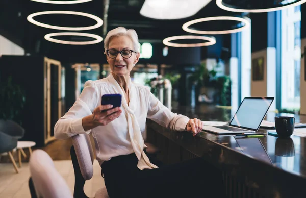 Positive smiling elderly female entrepreneur sitting at table with laptop in meeting room and browsing mobile phone while looking at screen and having free time after busy office schedule