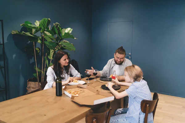 From above of content family in casual clothes sitting at wooden table with pizza and beverages while having dinner together in modern apartment